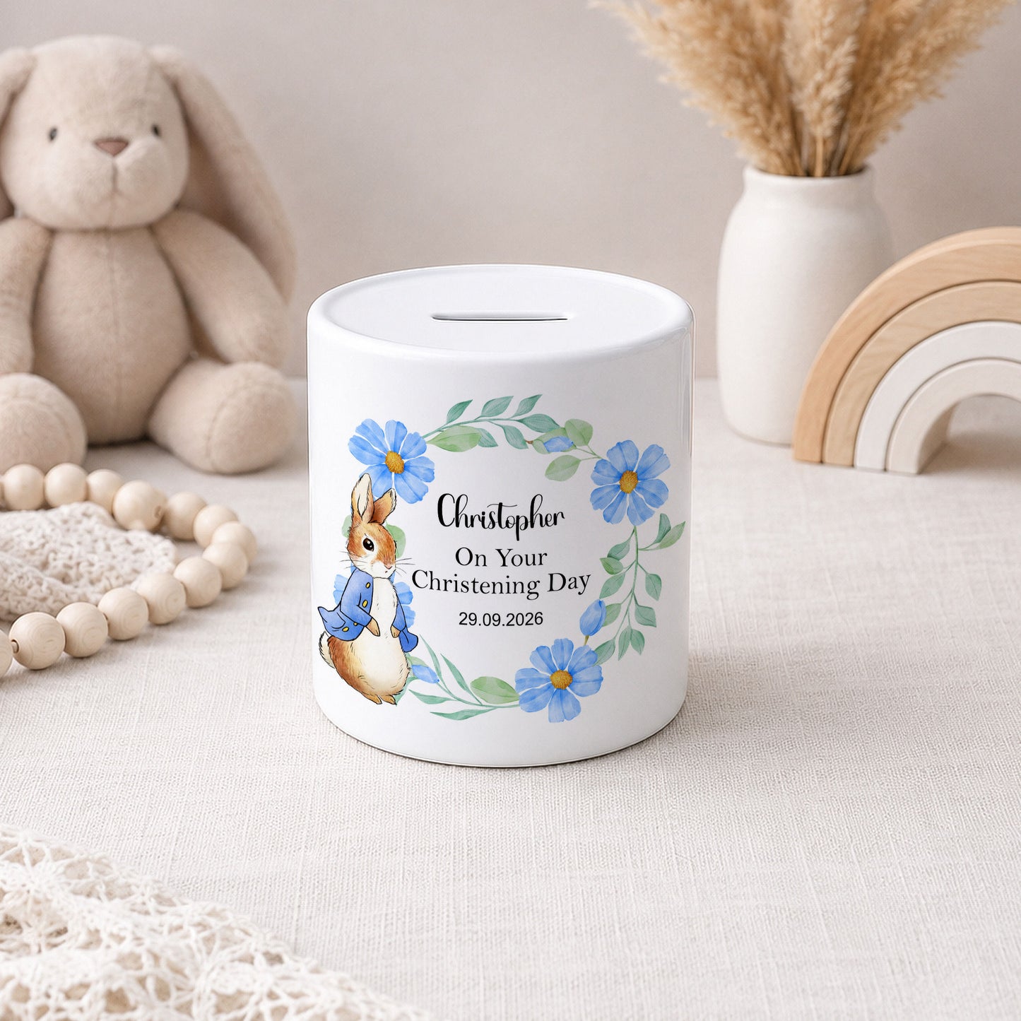 A white ceramic piggy bank with a personalised christening day design, featuring a watercolour rabbit in a blue outfit, blue flowers, and the name 'Christopher' on a light beige fabric surface with soft toys and decorative items in the background.