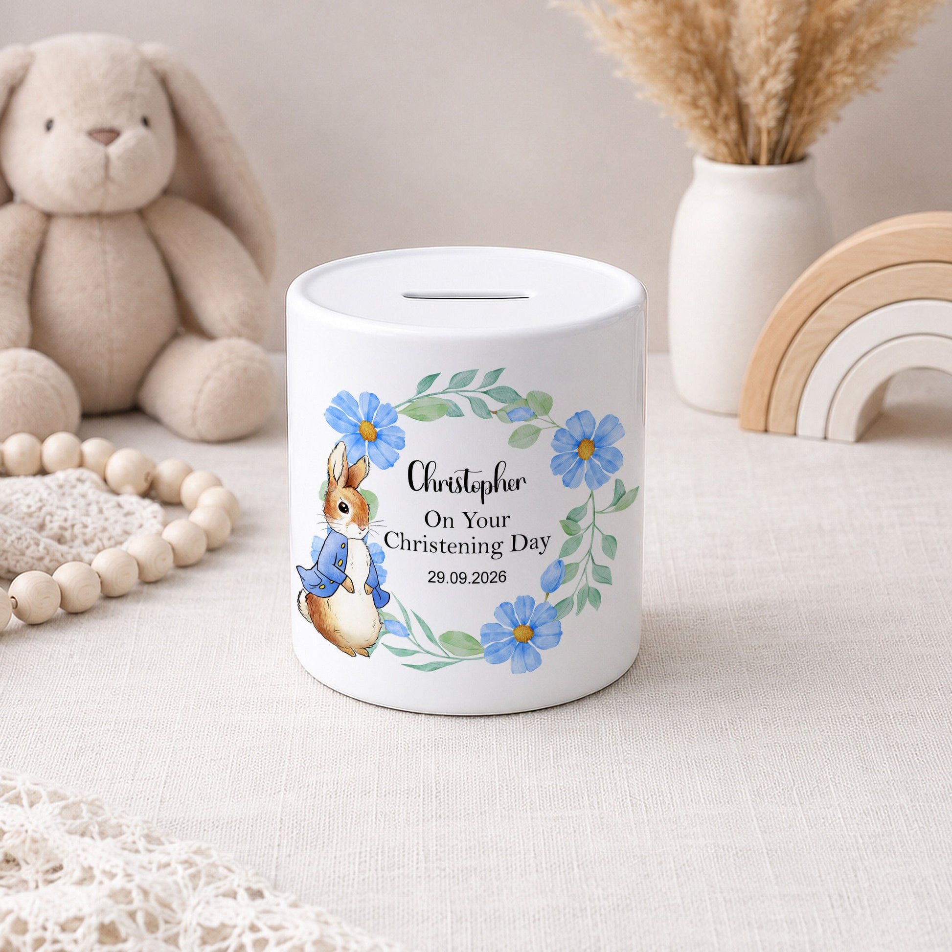 A white ceramic piggy bank with a personalised christening day design, featuring a watercolour rabbit in a blue outfit, blue flowers, and the name 'Christopher' on a light beige fabric surface with soft toys and decorative items in the background.