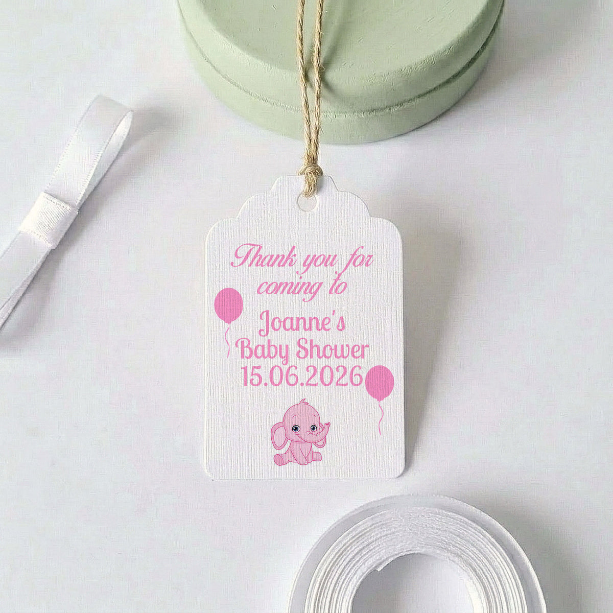 A personalised baby shower thank you gift tag with a pink elephant and balloon design, hanging from a twine string. The white textured tag features pink text reading 'Thank you for coming to Joanne's Baby Shower 15.