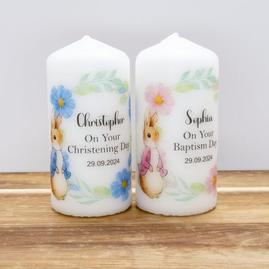 Two personalised white candles standing side by side on a wooden surface against a light grey background. Each candle features a watercolour-style beaver illustration, floral accents, and printed text reading 'On Your [Christening/Baptism Day]' with a date.