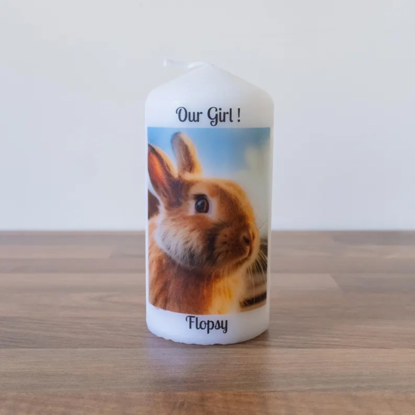 A white pillar candle standing on a wooden surface against a plain light wall. The candle features a printed photo of a brown rabbit with the text 'Our Girl!' at the top and the name 'Flopsy' at the bottom.