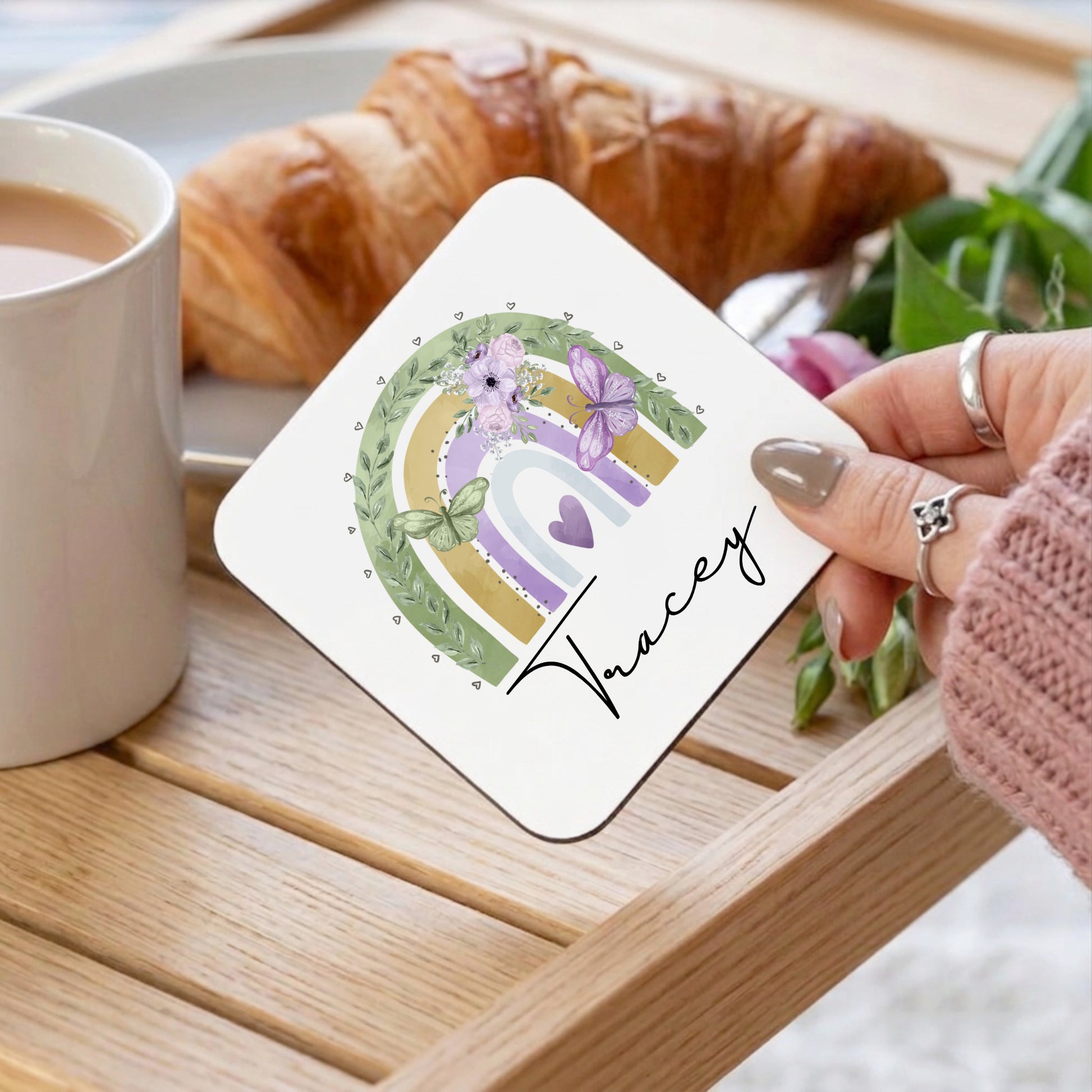 Coaster with floral design and name 'Tracey' held by a hand on a table with coffee and croissant.