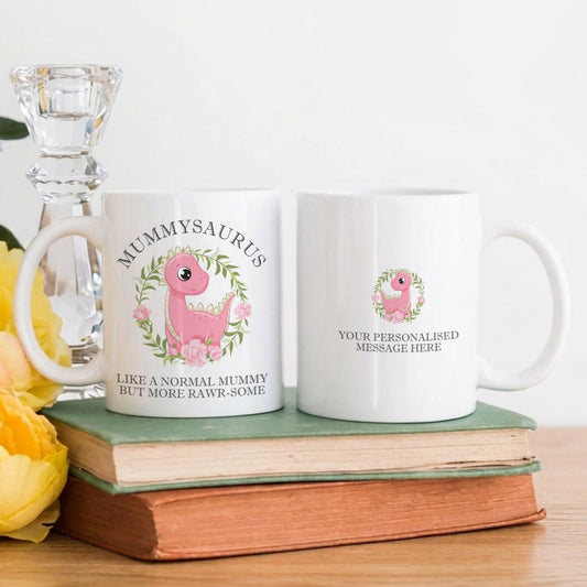 Two mugs with pink dinosaur designs on a wooden surface with flowers and a candle.