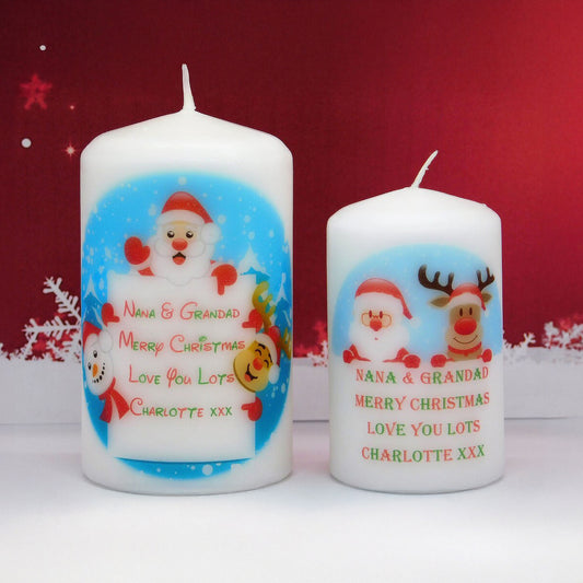 Two personalised white Christmas pillar candles with Santa and reindeer illustrations, shown on a white surface against a red background with snowflake decorations.