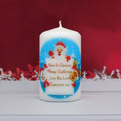 A white pillar candle with a personalised Christmas design featuring Santa Claus and a reindeer, with the text 'NANA & GRANDAD MERRY CHRISTMAS LOVE YOU LOTS CHARLOTTE XXX'. The candle is set against a dark red background with white decorative snowflakes.