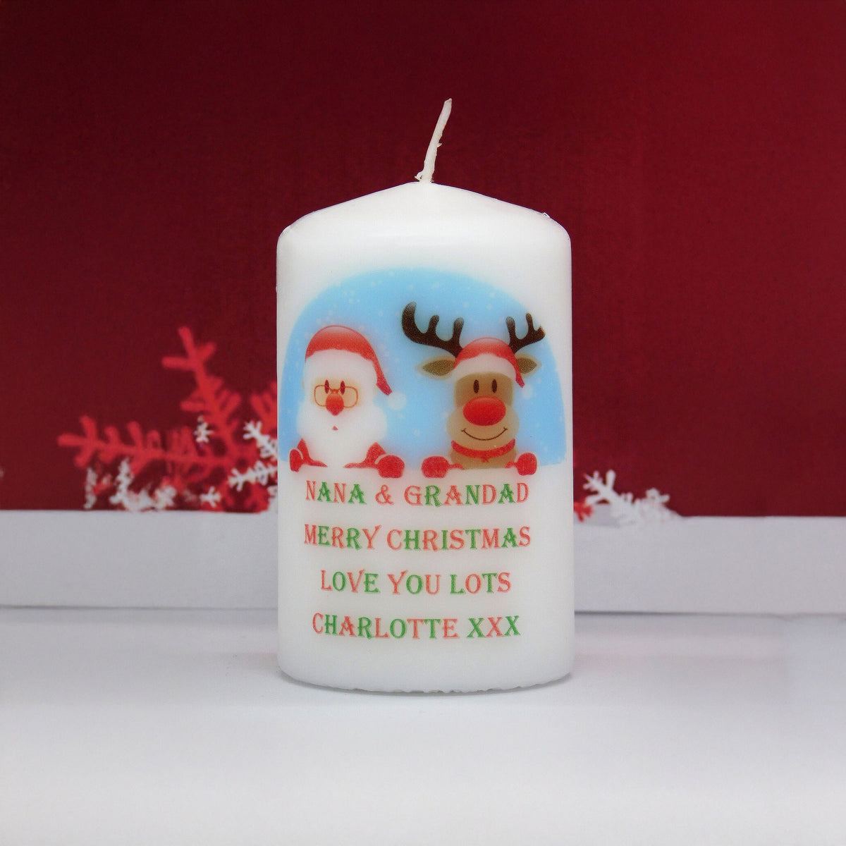 A white pillar candle with a personalised Christmas design featuring Santa Claus and a reindeer, with the text 'NANA & GRANDAD MERRY CHRISTMAS LOVE YOU LOTS CHARLOTTE XXX'. The candle is set against a dark red background with white decorative snowflakes.
