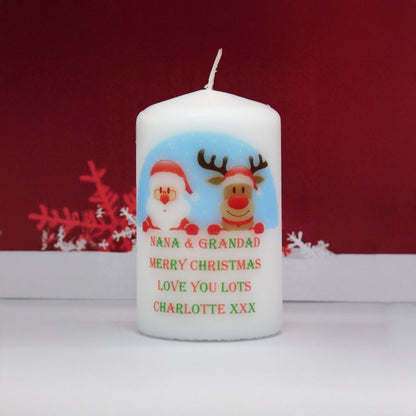 A white pillar candle with a personalised Christmas design featuring Santa Claus and a reindeer, with the text 'NANA & GRANDAD MERRY CHRISTMAS LOVE YOU LOTS CHARLOTTE XXX'. The candle is set against a dark red background with white decorative snowflakes.