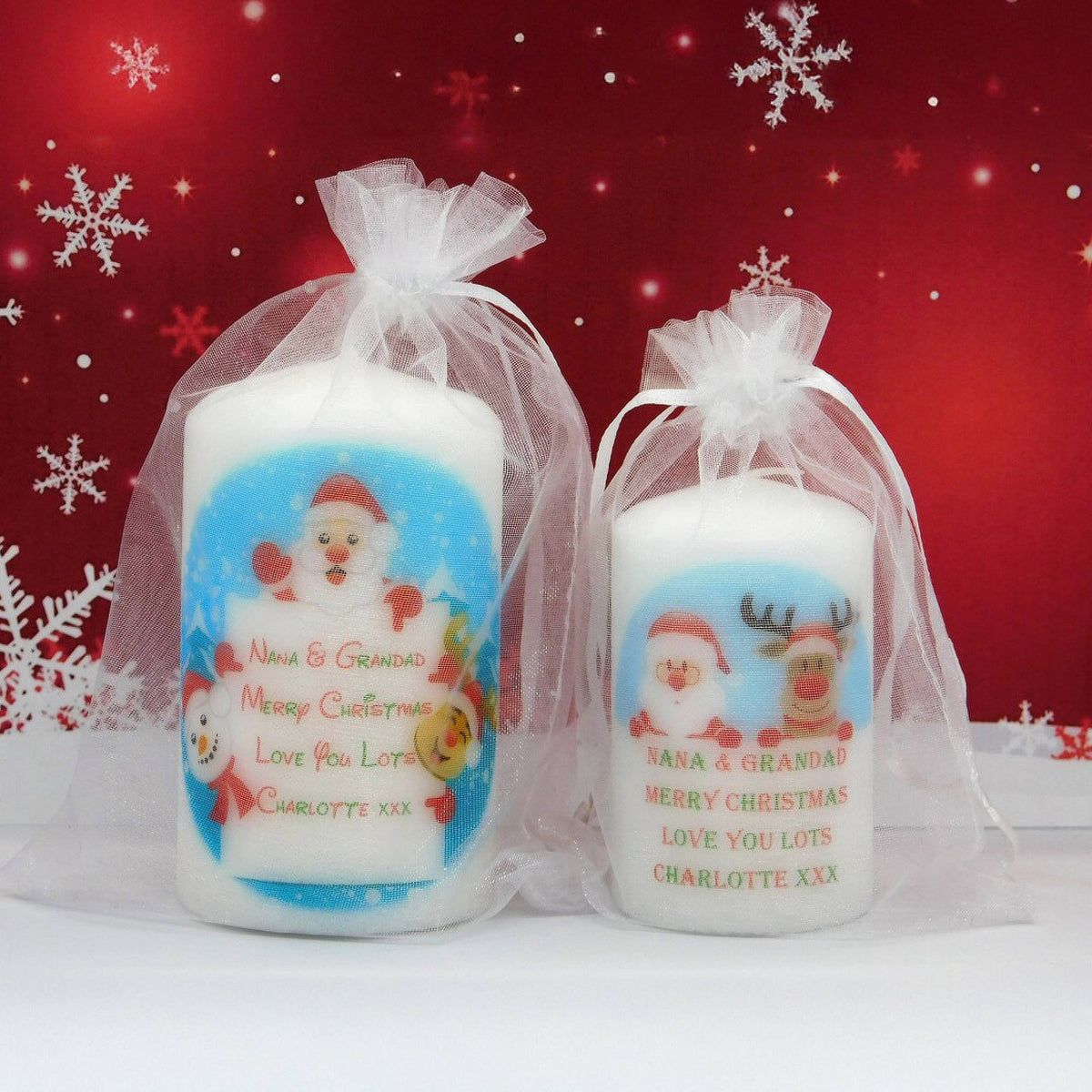 Two personalised white Christmas pillar candles in clear tulle gift bags, featuring Santa and reindeer illustrations with the text 'Nana & Grandpa Merry Christmas Love You Lots Charlotte, XXX' against a red holiday background with snowflakes.