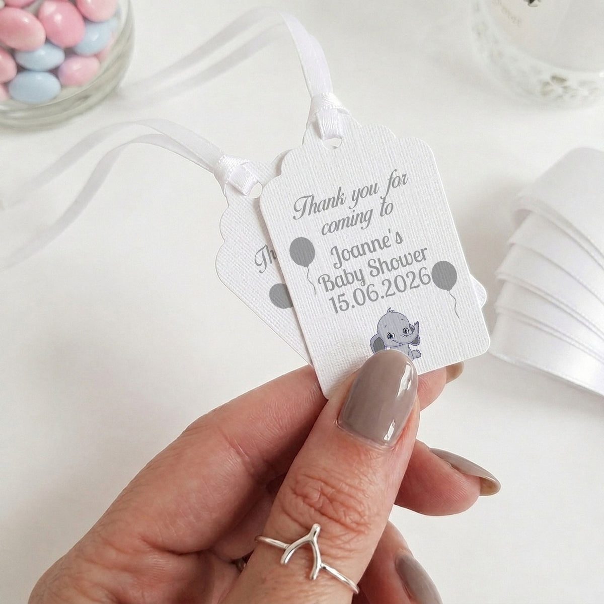 A hand with taupe nail polish holds two white personalised baby shower favour tags with white ribbons. The tags feature grey text reading 'Thank you for coming to Joanne's Baby Shower 15.