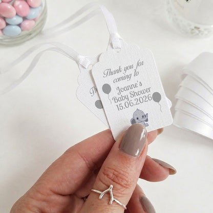 A hand with taupe nail polish holds two white personalised baby shower favour tags with white ribbons. The tags feature grey text reading 'Thank you for coming to Joanne's Baby Shower 15.