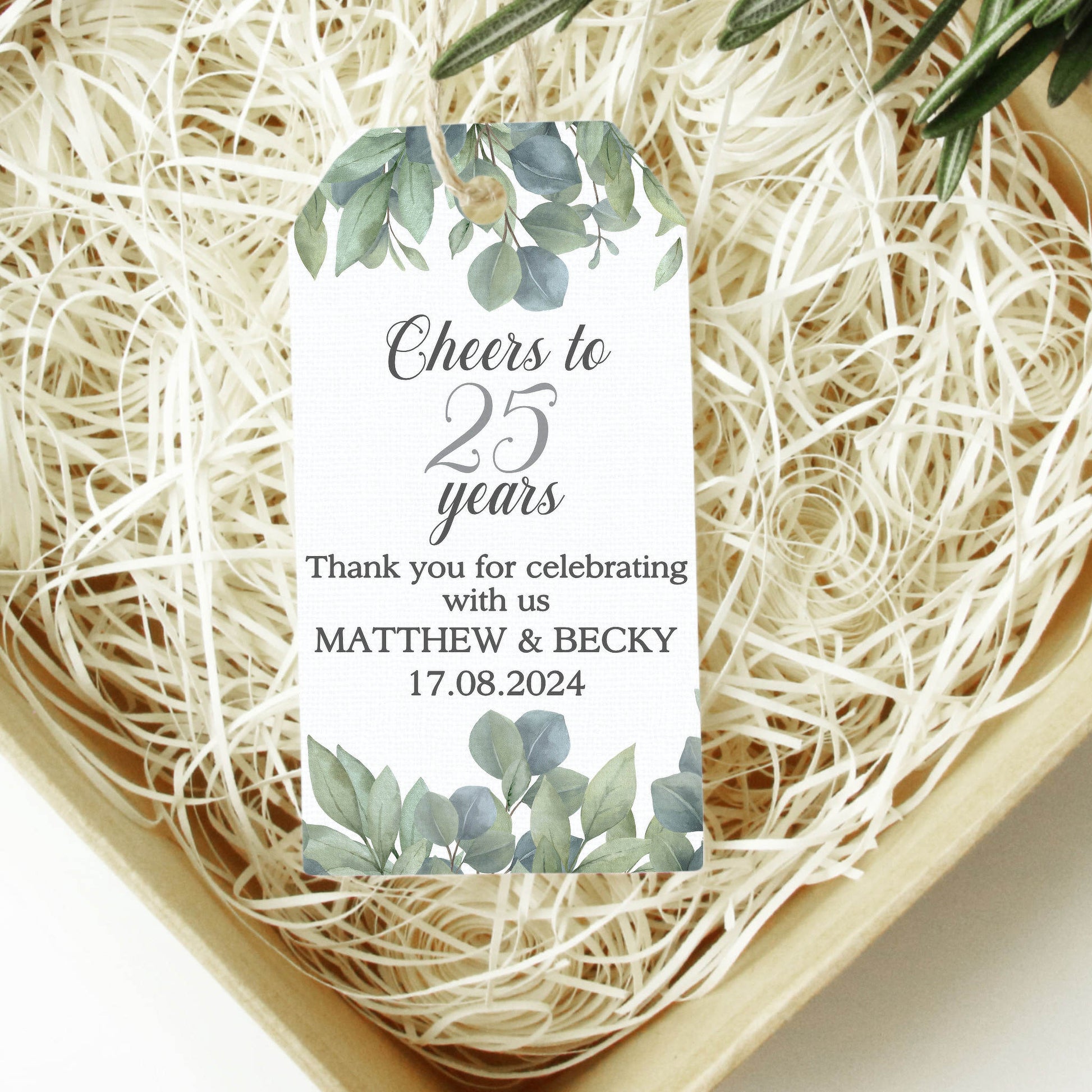 A rectangular white thank-you card with green eucalyptus illustrations at the top and bottom, placed on a bed of shredded paper inside a light wood box. The card reads, 'Cheers to 25 years Thank you for celebrating with us MATTHEW & BECKY.