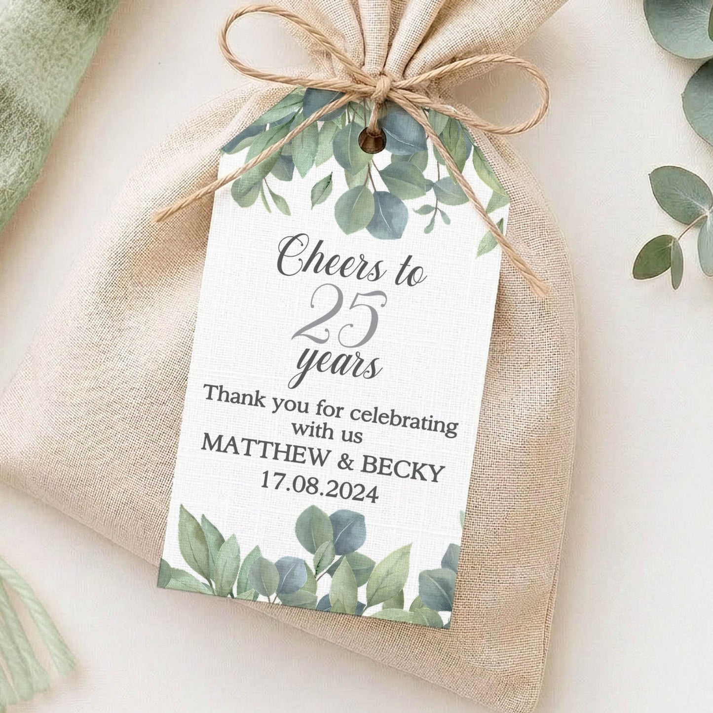 A small beige fabric pouch tied with twine and decorated with a custom printed tag featuring green watercolour leaves, styled on a light surface with a mint throw and eucalyptus branches.