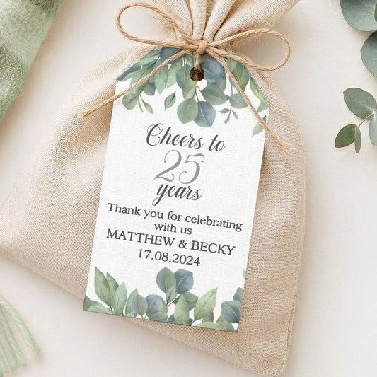 A small beige fabric pouch tied with twine and decorated with a custom printed tag featuring green watercolour leaves, styled on a light surface with a mint throw and eucalyptus branches.