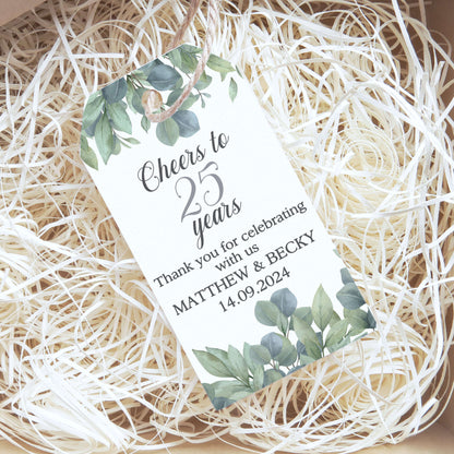 A personalised 25th birthday gift tag with green watercolour eucalyptus leaves and a small bird illustration, resting on white shredded paper inside a kraft gift box.