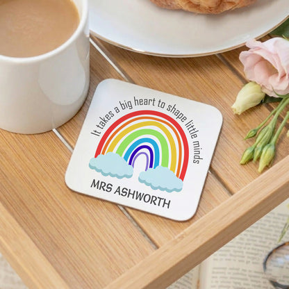 Coaster with rainbow design and text on a wooden table with a mug and flowers.