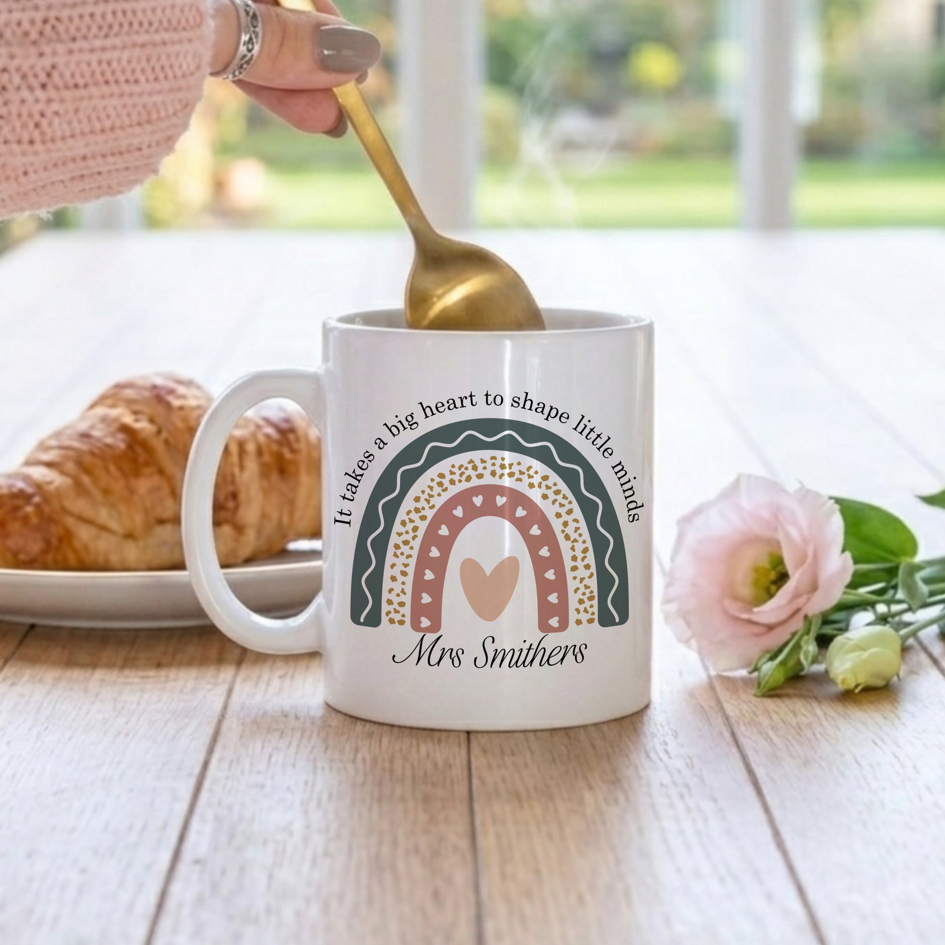 White mug with rainbow design and text, held by a hand with a gold spoon, on a wooden table with bread and flowers.