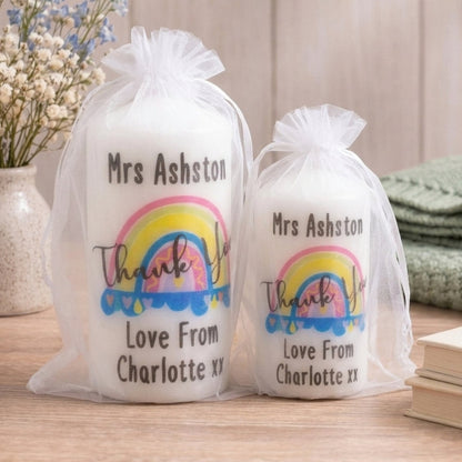 Two white pillar candles in sheer white organza bags displayed side by side on a wooden board against a light grey background, showing both available sizes. Each candle is printed with a colourful hand-drawn rainbow illustration in pink, yellow and blue, with the text 'Thank You' in script font, personalised with the name at the top and 'Love From Charlotte xx' at the bottom. The larger 120mm candle is on the left and the smaller 100mm candle is on the right. Personalised rainbow thank you teacher