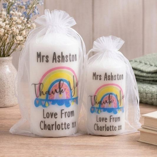 Two white pillar candles in sheer white organza bags displayed side by side on a wooden board against a light grey background, showing both available sizes. Each candle is printed with a colourful hand-drawn rainbow illustration in pink, yellow and blue, with the text 'Thank You' in script font, personalised with the name at the top and 'Love From Charlotte xx' at the bottom. The larger 120mm candle is on the left and the smaller 100mm candle is on the right. Personalised rainbow thank you teacher