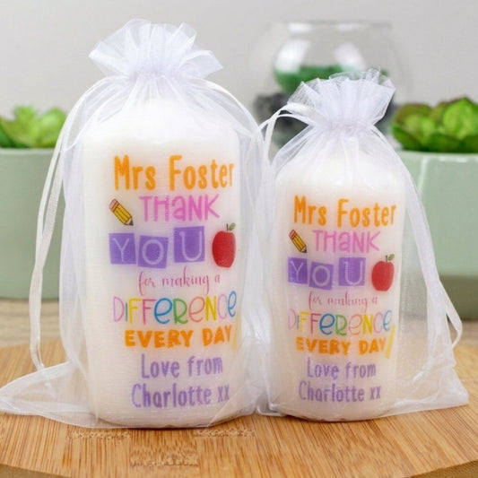 Two personalised candles with printed text 'Mrs Foster THANK YOU for making a DIFFERENCE every day' with additional personalized text that reads 'Love from Charlotte xx'. The candles are placed in transparent packaging.