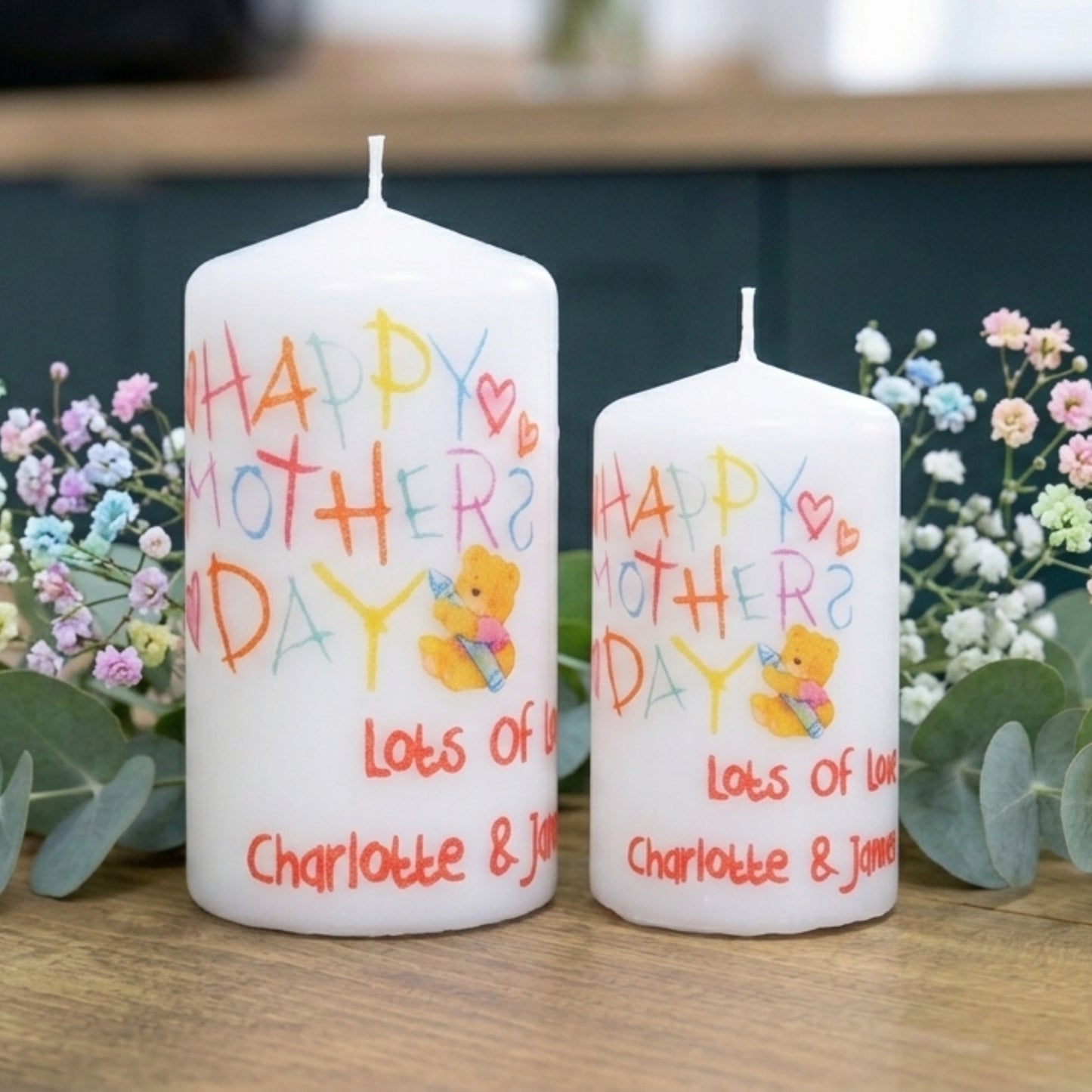 Two white candles with 'Happy Mother's Day' text and a teddy bear illustration, surrounded by flowers on a wooden surface.