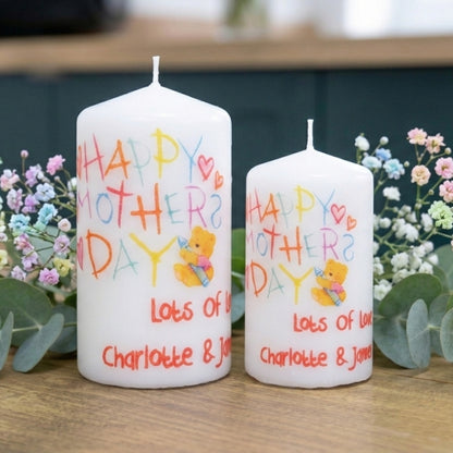 Two white candles with 'Happy Mother's Day' text and a teddy bear illustration, surrounded by flowers on a wooden surface.