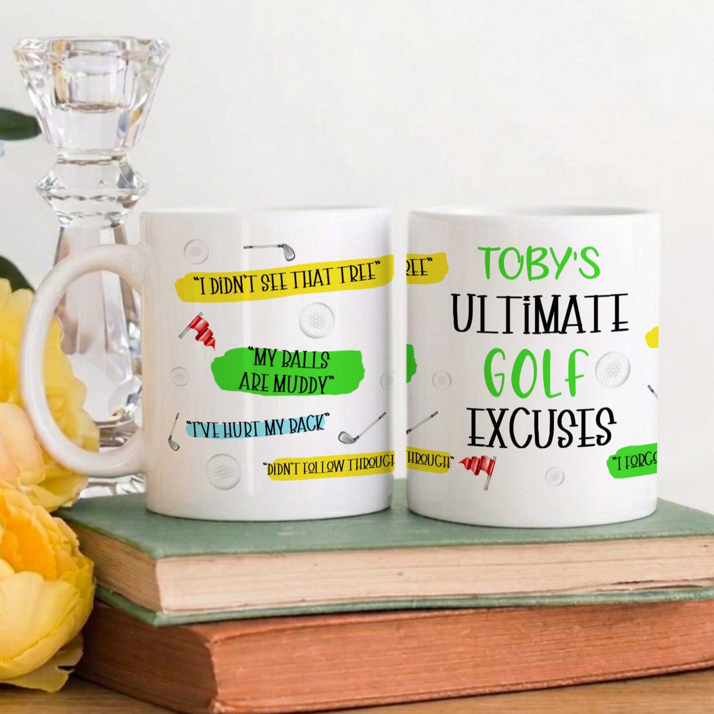 Two white ceramic mugs with golf-themed printed quotes and graphics, displayed on stacked vintage books next to yellow flowers and a glass candle holder against a plain white wall.