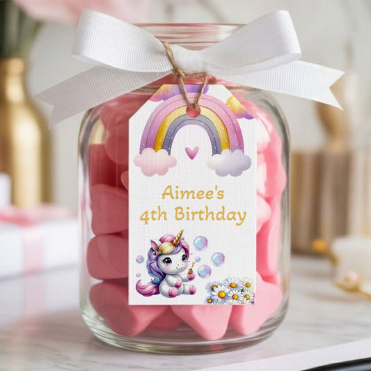 A clear glass jar filled with pink heart-shaped candies, tied with a white ribbon and a custom printed tag that reads 'Aimee's 4th Birthday' with a unicorn and rainbow illustration. The jar sits on a marble surface with blurred gift boxes and flowers in the background.