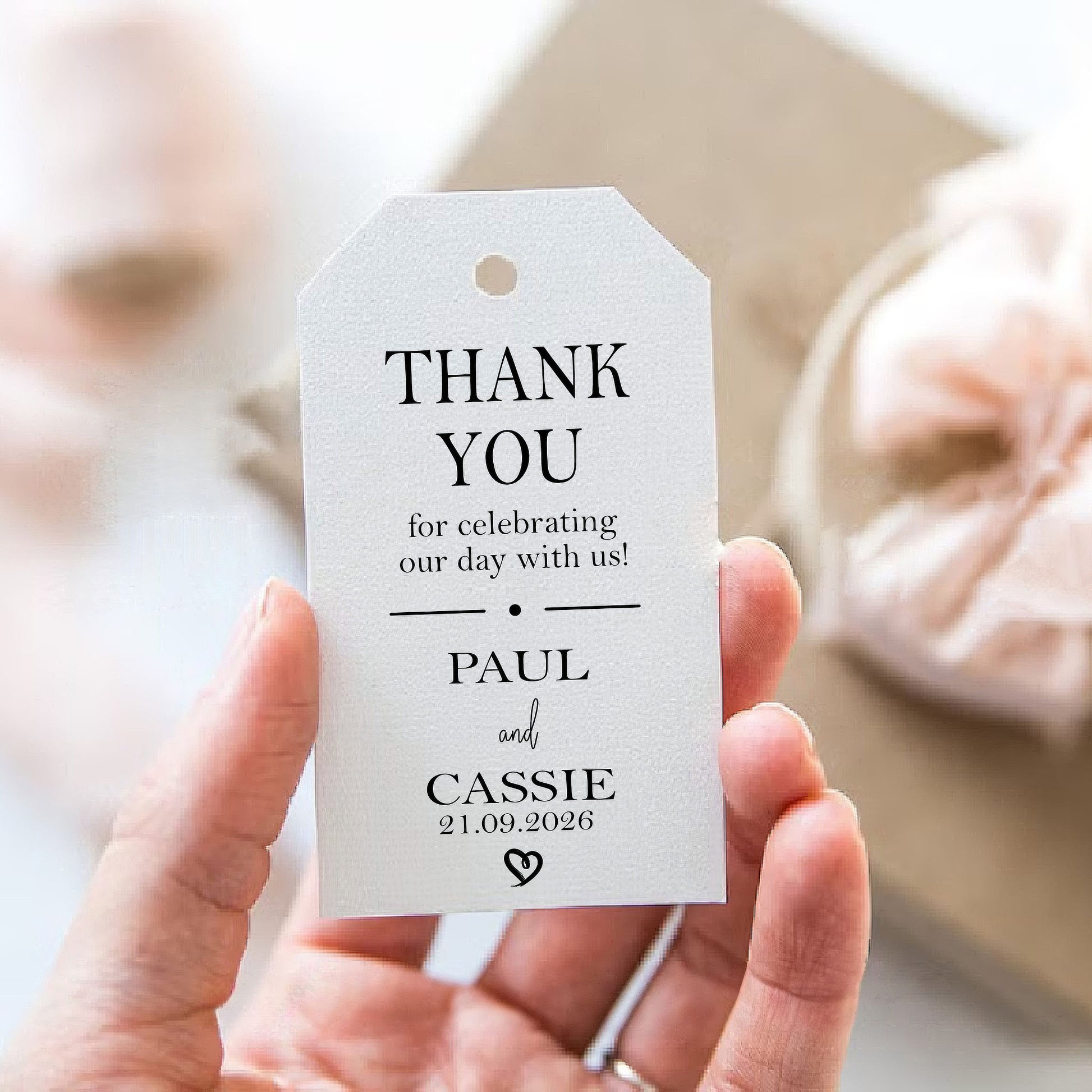 A hand holding a white personalised thank you gift tag with black text, set against a soft-focus background of a kraft paper bag and a pale pink fabric.