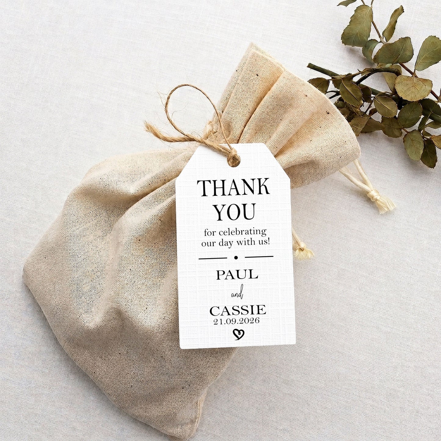 A small natural linen drawstring pouch with a white printed thank-you tag tied to it, photographed on a light neutral surface with eucalyptus leaves in the corner.