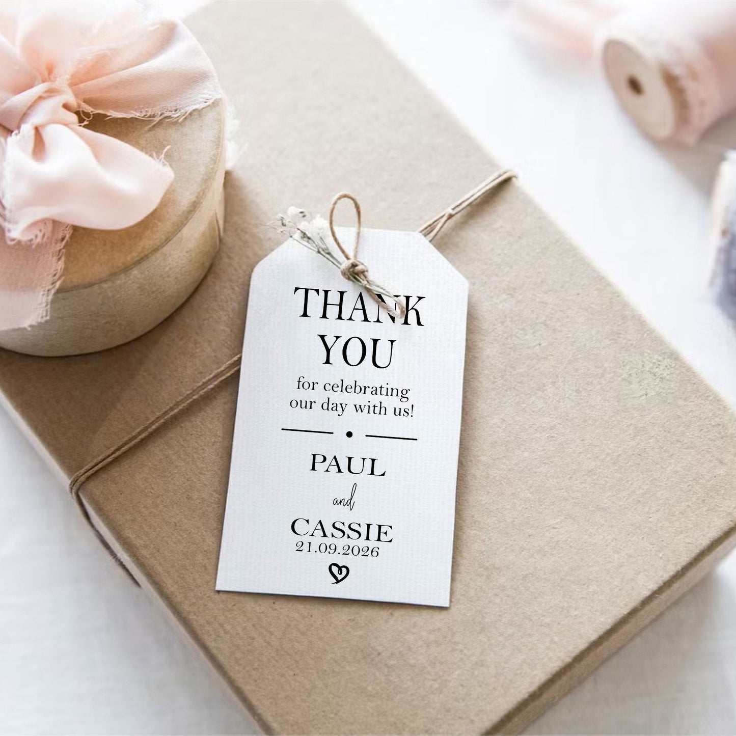 A white wedding thank-you gift tag with black text, tied to a kraft paper gift box with a pink ribbon bow.