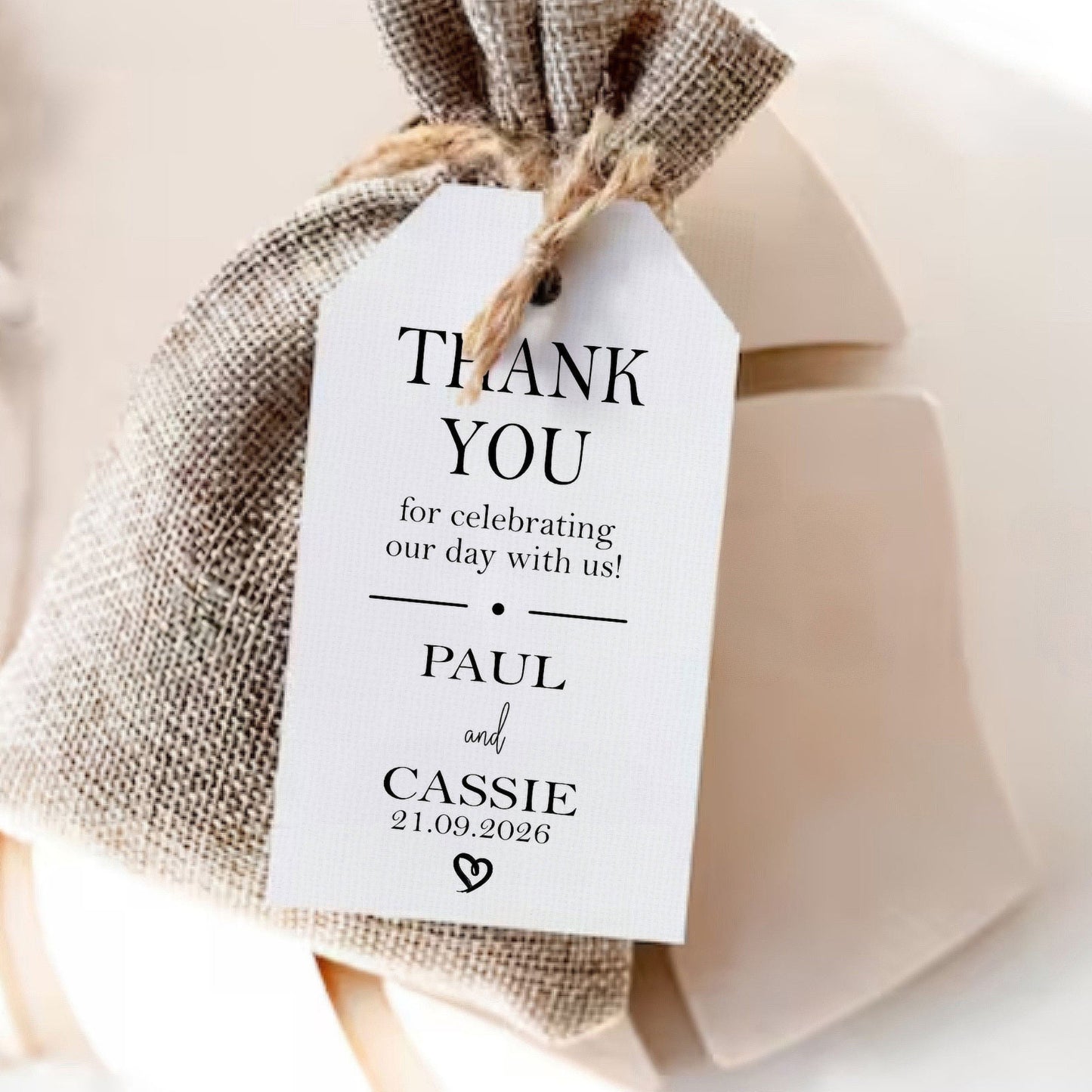 A small burlap drawstring gift bag tied with twine and labelled 'THANK YOU' for celebrating with Paul and Cassie, resting on a white gift box.