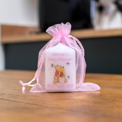 A white baby shower favour candle featuring a printed illustration of Winnie the Pooh and the text 'Hannah's Baby Shower 15th April 2026', tied in a sheer pink organza drawstring bag and resting on a wooden table.