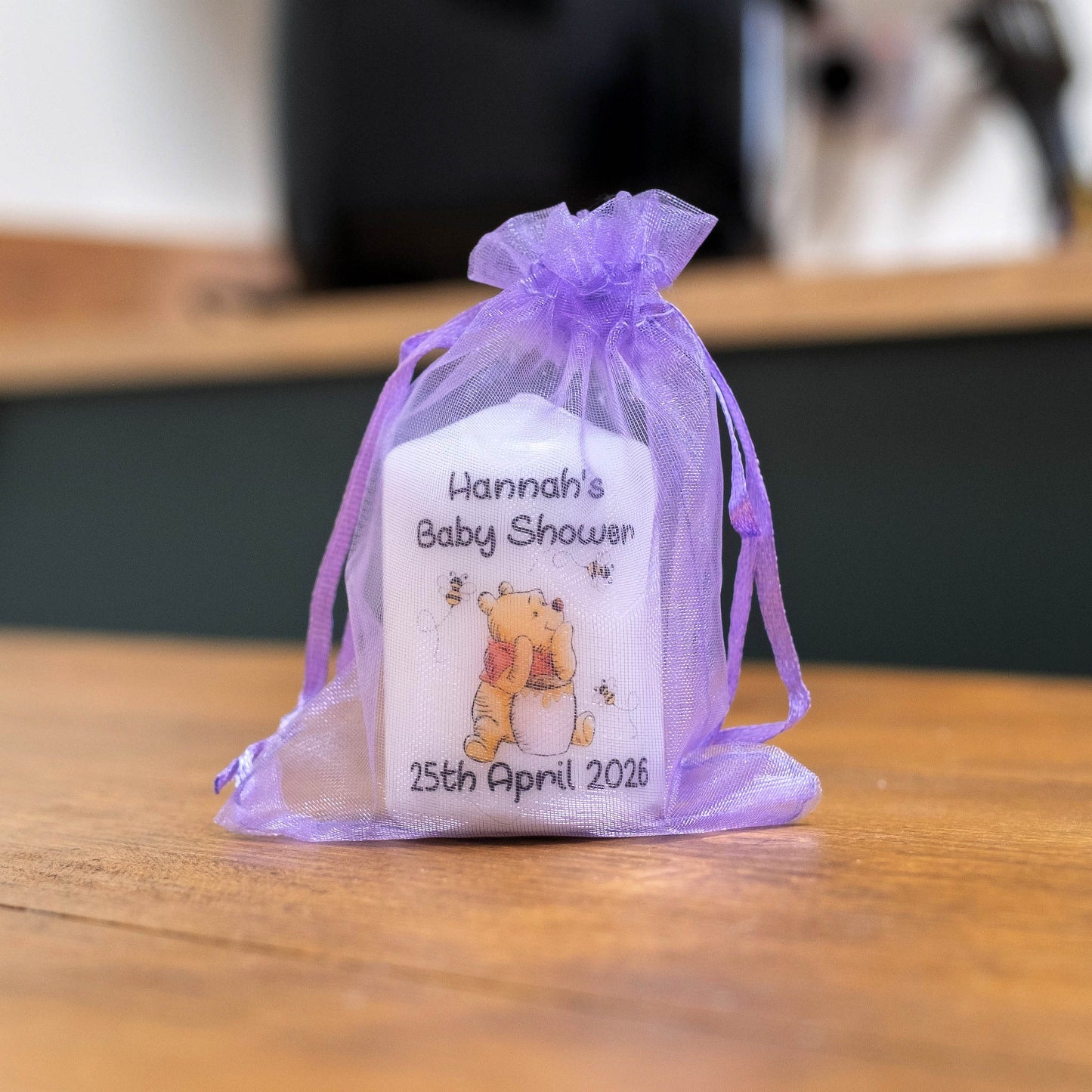 A white baby shower favour candle tied with a sheer lavender purple organza drawstring, featuring a custom printed design with the text 'Hannah's Baby Shower' and a Winnie the Pooh illustration, sitting on a wooden table.