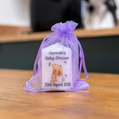 A white baby shower favour candle tied with a sheer lavender purple organza drawstring, featuring a custom printed design with the text 'Hannah's Baby Shower' and a Winnie the Pooh illustration, sitting on a wooden table.