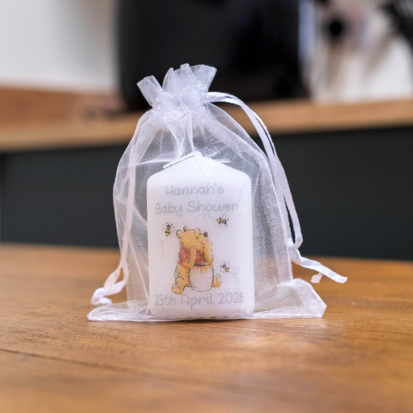 A white baby shower favour candle with a Winnie the Pooh bear illustration and personalised text, tied inside a sheer white organza drawstring pouch on a wooden table.