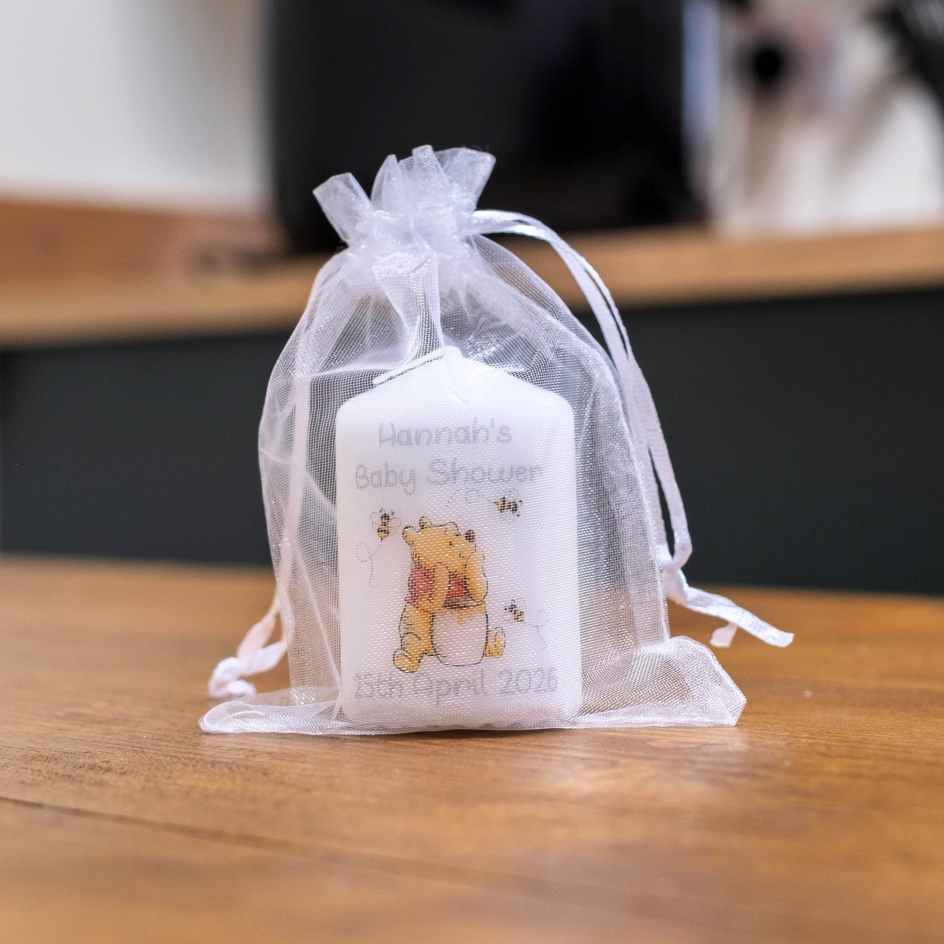 A white baby shower favour candle with a Winnie the Pooh bear illustration and personalised text, tied inside a sheer white organza drawstring pouch on a wooden table.