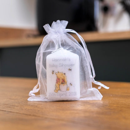 A white baby shower favour candle with a Winnie the Pooh bear illustration and personalised text, tied inside a sheer white organza drawstring pouch on a wooden table.