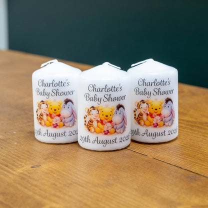 Three white candles on a wooden table, printed with the text 'Charlotte's Baby Shower' and a colourful illustration of Winnie the Pooh characters, with the date '19th August 2023' at the bottom.