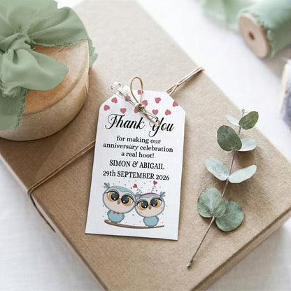 A personalised thank you gift tag with owl illustrations and heart accents, wrapped around a brown kraft paper gift box with twine. The scene includes a mint green ribbon on a wrapped package, a sprig of eucalyptus, and soft-focus decorative elements in the background.