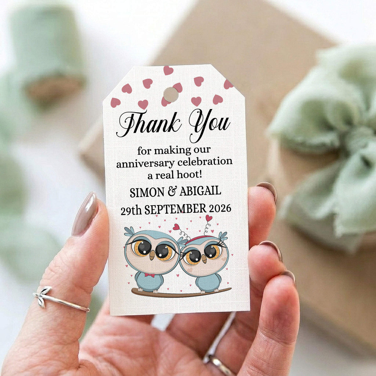 A hand holds a personalised thank you gift tag with a cute owl illustration and pink hearts. The tag reads: 'Thank You for making our anniversary celebration a real hoot! SIMON & ABIGAIL 29th SEPTEMBER 2026'.