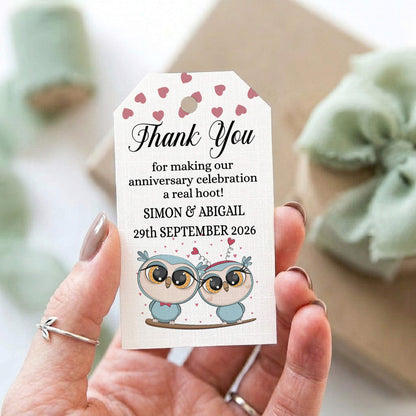 A hand holds a personalised thank you gift tag with a cute owl illustration and pink hearts. The tag reads: 'Thank You for making our anniversary celebration a real hoot! SIMON & ABIGAIL 29th SEPTEMBER 2026'.