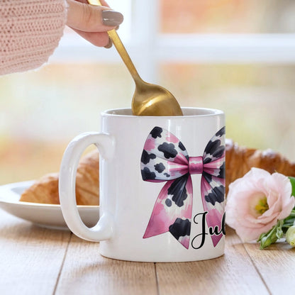Personalised 'Julie' mug with a pink and black cow print bow design, being stirred with a gold spoon alongside a croissant and pink flower