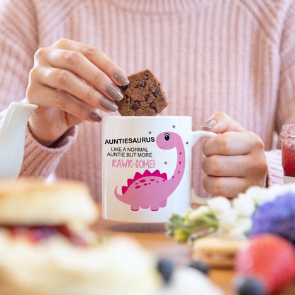 A person in a pink ribbed sweater holds a white ceramic mug with a pink dinosaur graphic and the text 'AUNTIESAURUS LIKE A NORMAL AUNTIE BUT MORE RAWR-ROME!' while placing a chocolate chip cookie into the mug. The background is a softly blurred breakfast table setting.