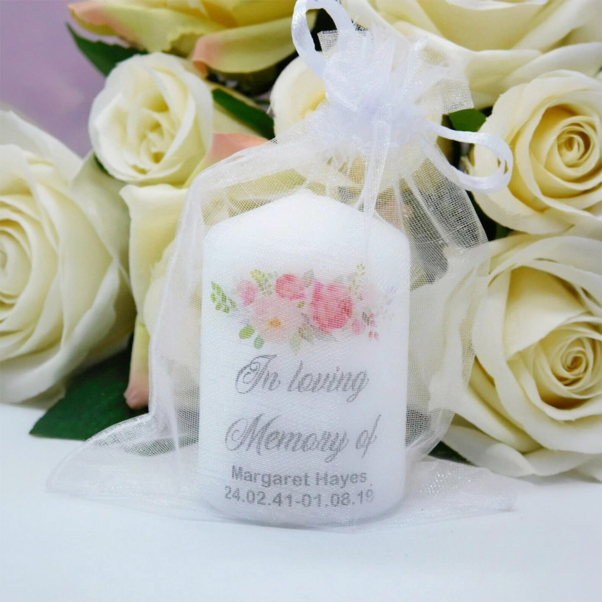A white memorial candle sealed in a sheer white organza bag, decorated with pink and green floral artwork and the printed text 'In Loving Memory of Margaret Hayes' and a date. The bag is styled in front of a bouquet of cream-coloured roses.