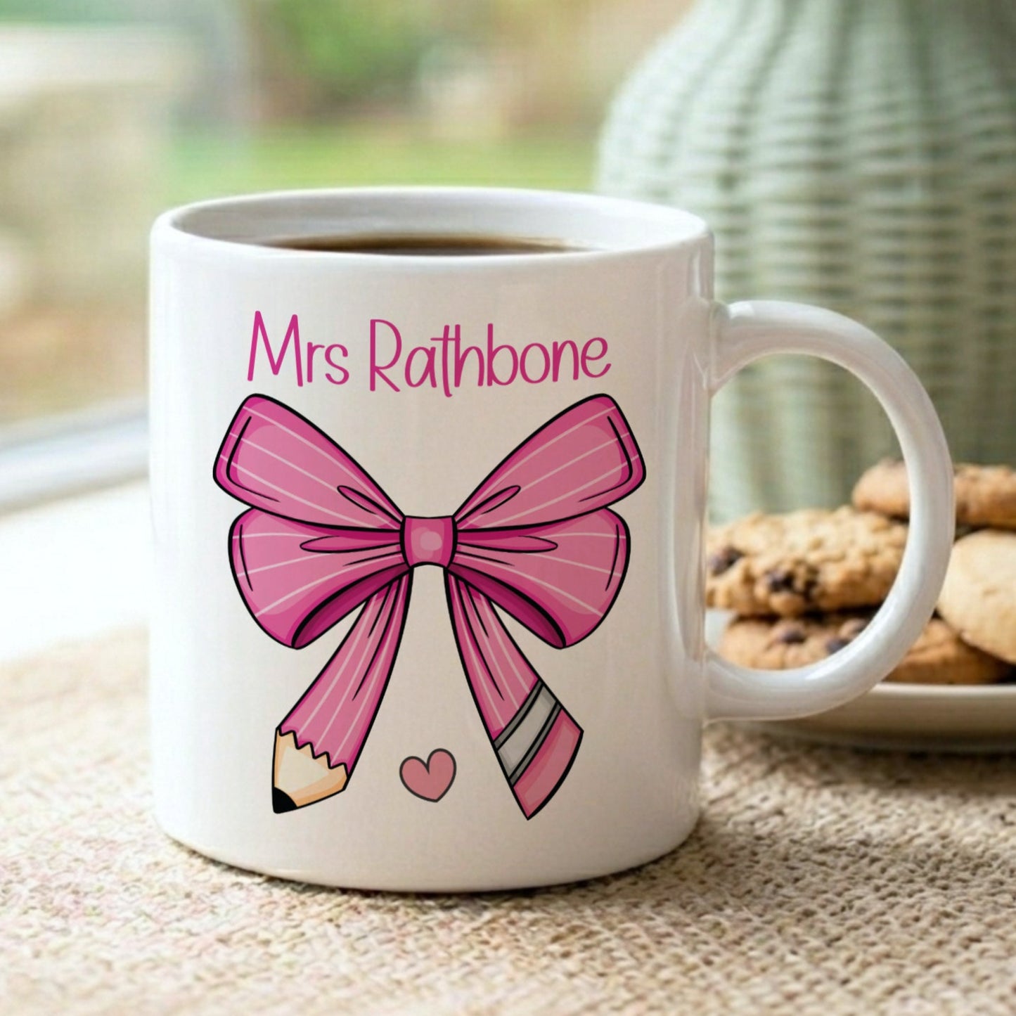 Personalised 'Mrs Rathbone' teacher mug with a pink bow and pencil design, displayed on a windowsill with cookies and a sage green vase