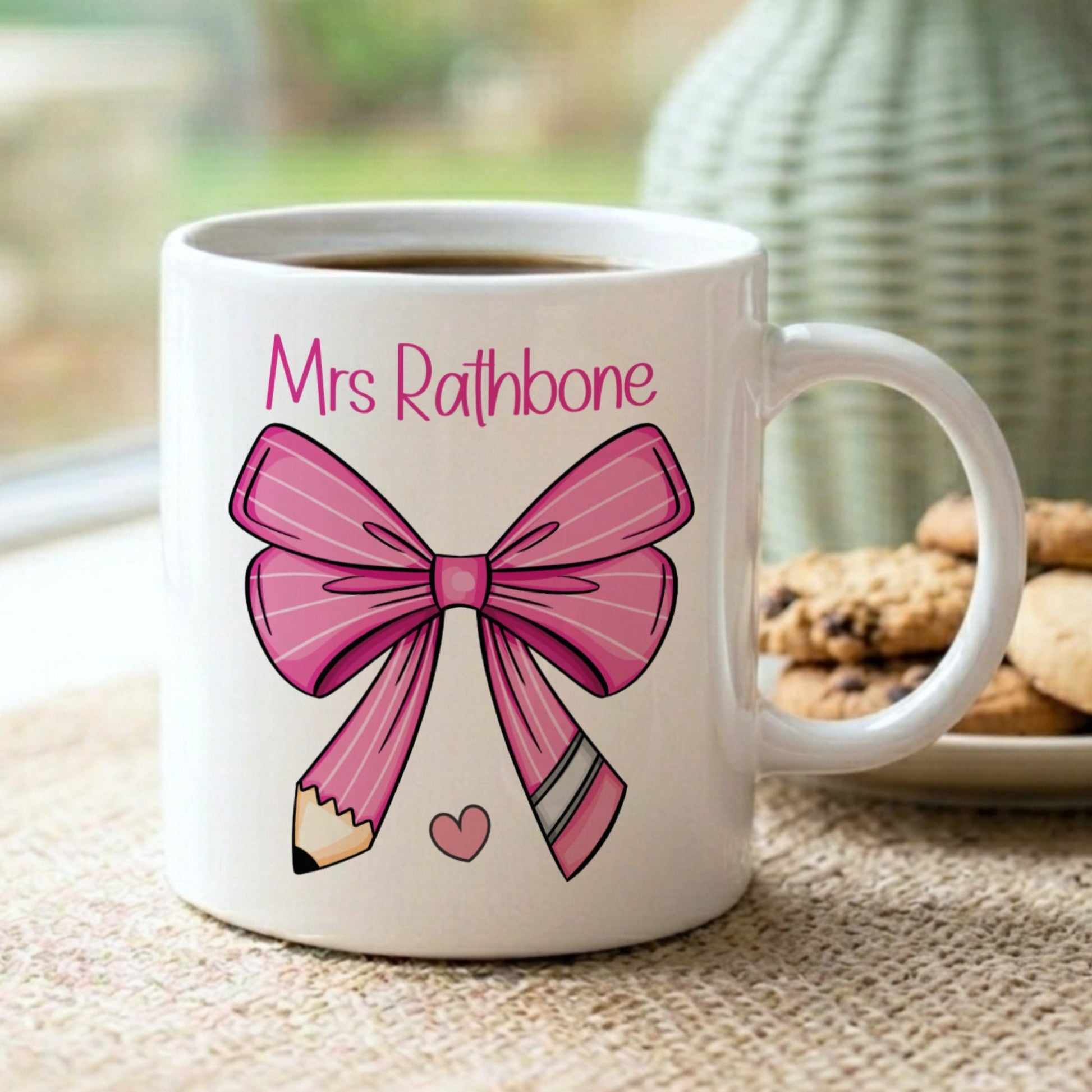 Personalised 'Mrs Rathbone' teacher mug with a pink bow and pencil design, displayed on a windowsill with cookies and a sage green vase