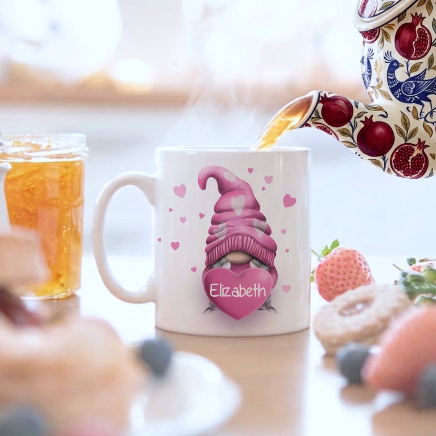 White ceramic mug with a pink gnome design holding a heart with the name "Elizabeth". The mug is decorated with pink hearts and is being filled with tea from a patterned teapot. A jar of jam and pastries are also visible.