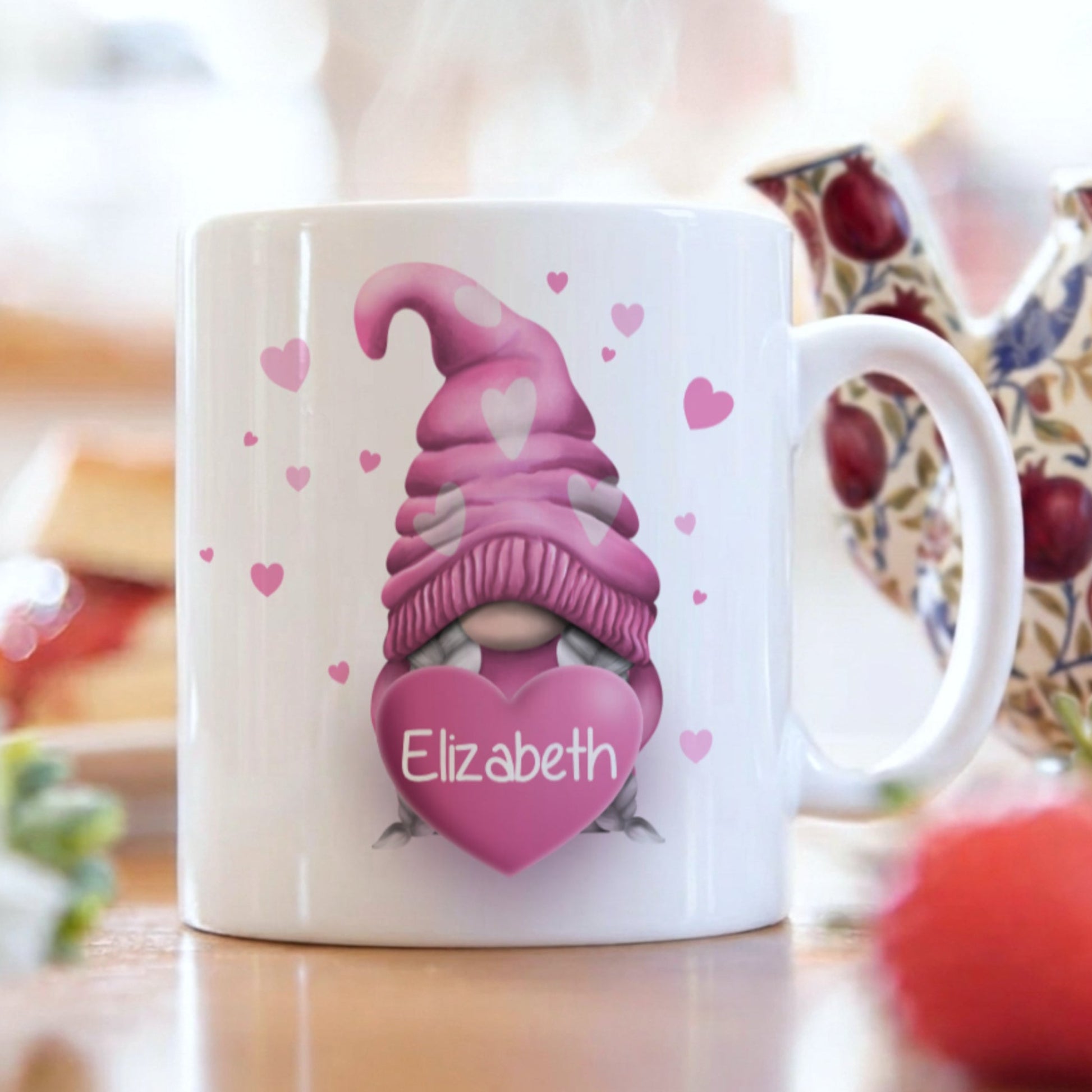 White ceramic mug with a pink gnome design holding a heart with the name "Elizabeth". The gnome has a pink hat with white hearts. Small pink hearts are scattered around the mug. A teapot and strawberries are in the background.