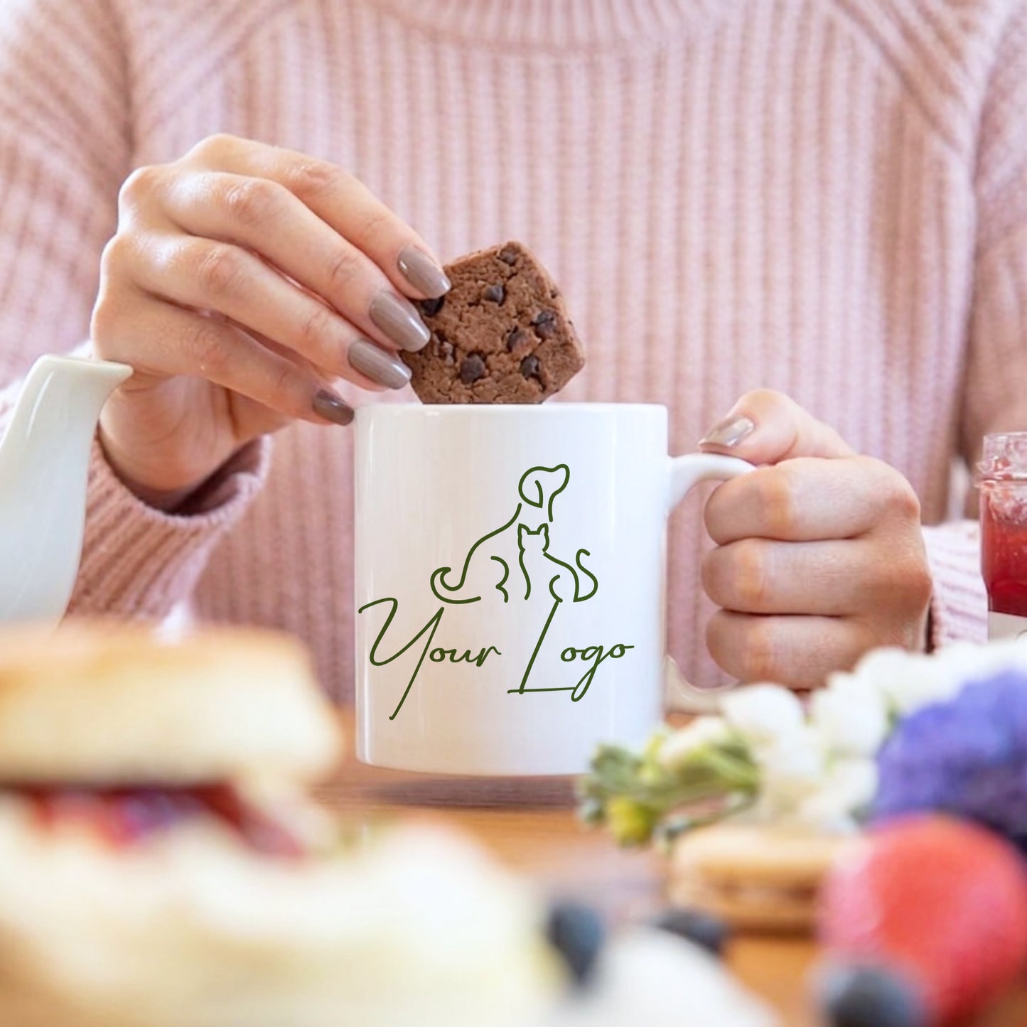 Person holding a mug with a logo and a cookie, blurred background