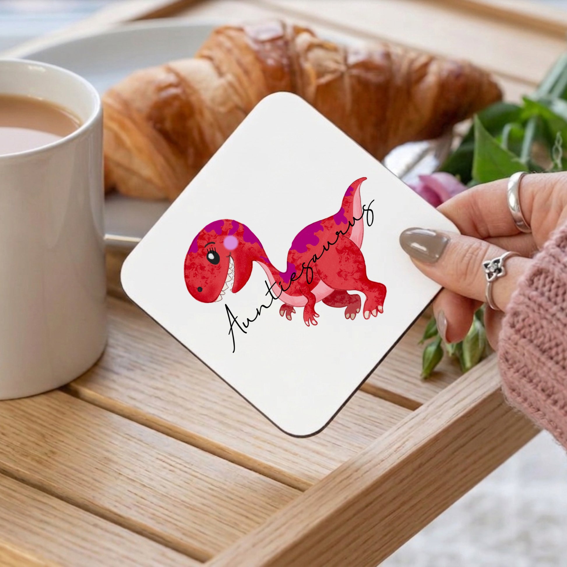 Coaster with dinosaur design held by a hand on a table with coffee and croissant
